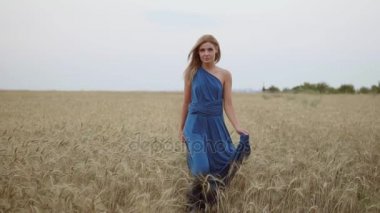 Kameraya bakarak altın buğday alan yürürken uzun mavi elbiseli güzel genç mutlu kız. Özgürlük kavramı. Slowmotion atış