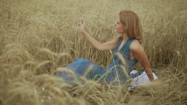 Attraente giovane donna seduta nel campo di grano dorato. La sua mano sta toccando le spighe. Concetto di raccolta. Raccolta. Colpo al rallentatore — Video Stock