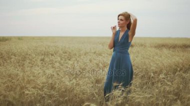 Yakışıklı genç kadın uzun mavi elbise ayakta buğday kök çalışırken, saçları çok dokunaklı ve doğanın keyfini altın buğday alanında. Özgürlük kavramı. Slowmotion atış