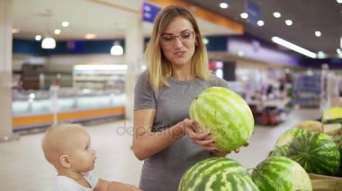 Genç anne bir karpuz, bir alışveriş sepeti içinde oturan küçük çocuğunu için gösterilen seçmektir. Süpermarkette alışveriş aile