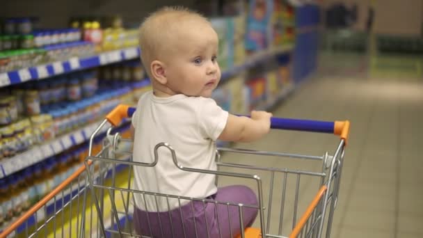 Малышка сидит в тележке с продуктами в супермаркете и ждёт, когда её мать вернётся с покупками. Семейный магазин с маленьким ребенком — стоковое видео