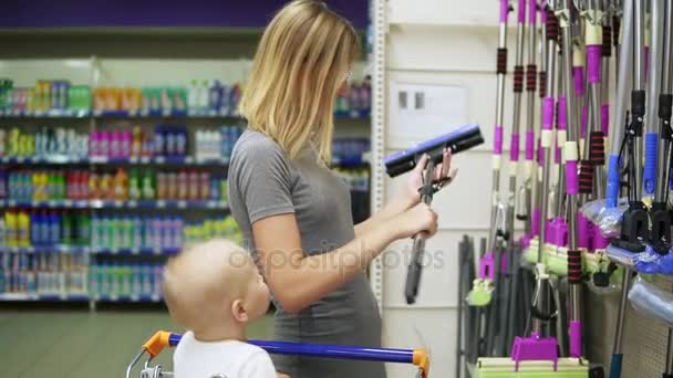 Joven madre atractiva es la elección de una fregona en el supermercado, mientras que su hijo pequeño está sentado en un carrito de comestibles. Compras familiares con un niño pequeño . — Vídeos de Stock