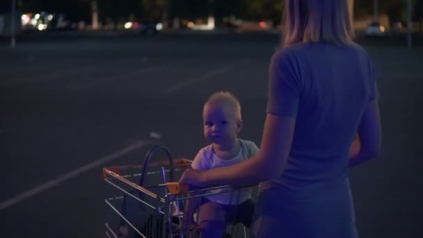 Pequeno bebê sentado em um carrinho de supermercado, enquanto sua mãe está empurrando o carrinho para a frente andando no lugar de estacionamento perto do supermercado depois de fazer compras à procura do carro — Vídeo de Stock