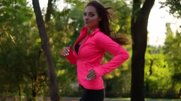 Eine junge Frau in leuchtend pinkfarbener Jacke läuft durch den sonnigen Stadtpark und übt im Freien. Schwebestativ stabilisiert Schuss, Zeitlupe. Morgentraining. Gesunder Lebensstil — Stockvideo