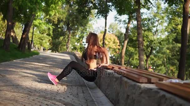 Молодая женщина в лифчике делает обратные отжимания в парке, слушая музыку. Слоумоушн — стоковое видео