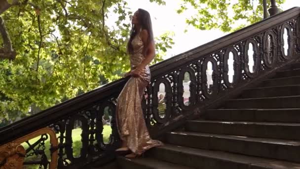Atractiva mujer en forma joven en un vestido largo brillante dorado camina por las escaleras en tacones altos en verano. Preparación de fiesta, modelo de moda. Disparo en cámara lenta — Vídeos de Stock