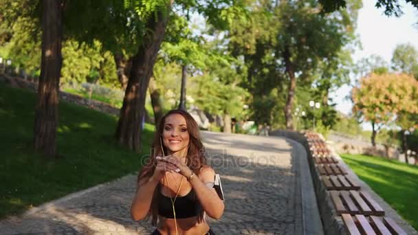 Mujer joven haciendo sentadillas en el parque escuchando la música, respirando lentamente y mirando a la cámara. Disparo en cámara lenta — Vídeo de stock