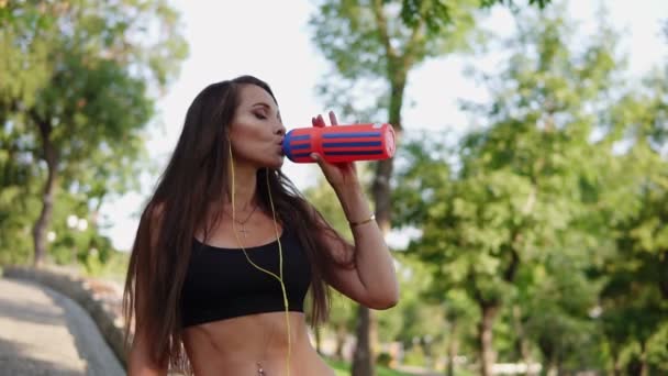 Jovem mulher fitness beber água de garrafa de plástico após jogging manhã, ouvindo a música e água potável da garrafa refrescando-se depois de correr no parque. Movimento lento — Vídeo de Stock