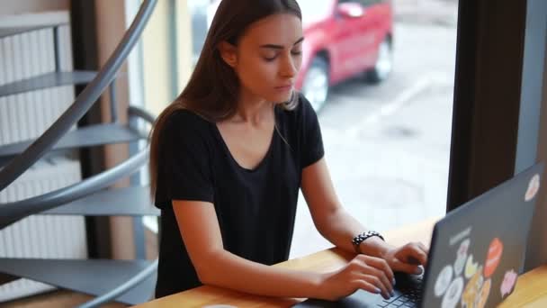 Mujer joven sentada en la cafetería junto a la ventana con su portátil y pensando qué escribir o cómo responder. Entonces ella tiene una idea y empieza a escribir — Vídeo de stock