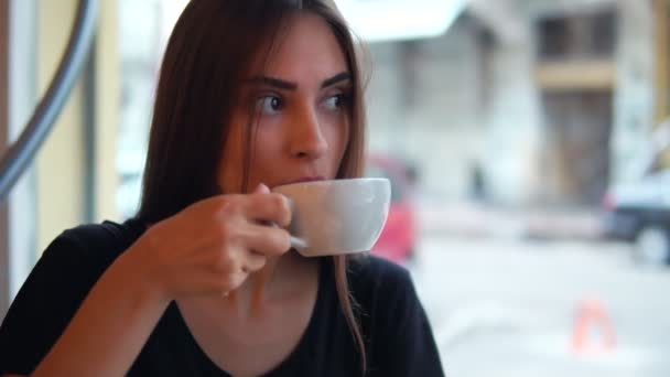 Close-up beeld van aantrekkelijke jonge vrouw dromen met een kop warme koffie zitten bij het raam in de coffeeshop. Breken na lang drukke dag. Ontspanning met een warme kop thee — Stockvideo
