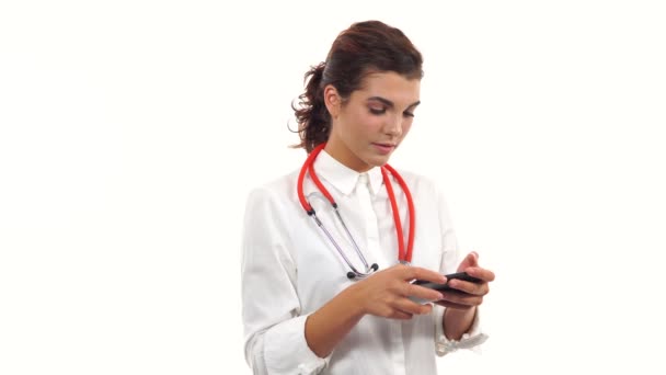 Retrato de una joven enfermera usando su smartphone para enviar mensajes de texto. Joven profesional médico con estetoscopio y bata de laboratorio aislada sobre fondo blanco — Vídeo de stock