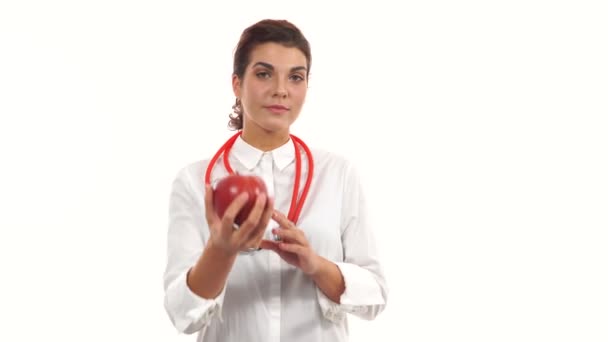 Jeune nutritionniste féminine amicale montrant une pomme rouge, conseillant une alimentation saine. Portrait de jeune professionnel avec stéthoscope et blouse isolé sur fond blanc — Video
