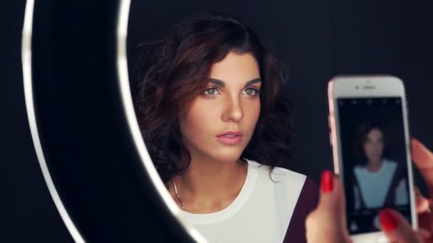 Close-up shot van jonge en mooie vrouw wordt gefotografeerd met mobiele telefoon vóór de ring-lamp. Aantrekkelijke model met natuurlijke make-up en bruine haren geïsoleerd op zwarte achtergrond — Stockvideo