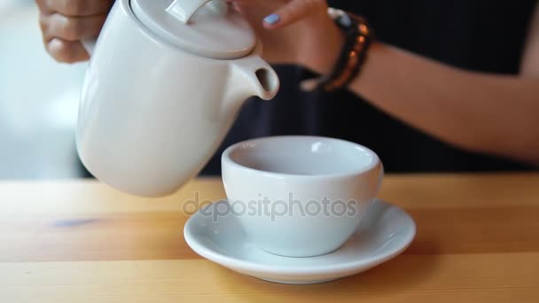 Vista de cerca de las manos femeninas vertiendo té caliente de la tetera en la taza de porcelana blanca en la mesa de madera en la cafetería — Vídeos de Stock