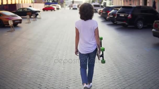 Vista posteriore di una donna che cammina nella vecchia strada della città tenendo il suo longboard al mattino, parcheggio in città. Bella vista sulla città vecchia — Video Stock