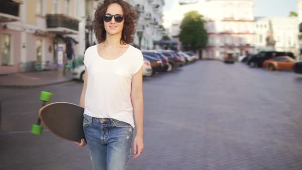 Gelukkige vrouw in zonnebril wandelen in de oude stad straat holding haar stijlvolle longboard in de ochtend. Slow-motion shot. Mooi uitzicht op de oude stad — Stockvideo