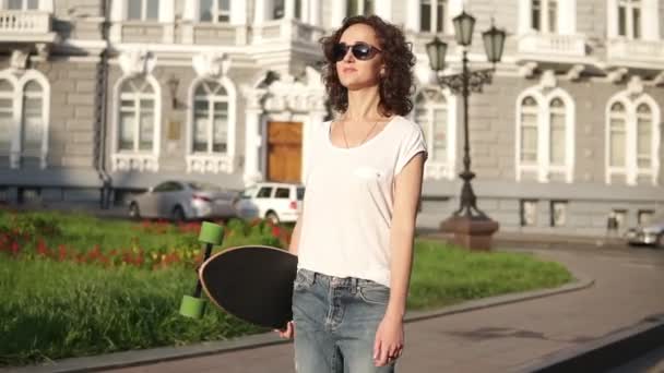 Belle jeune femme en lunettes de soleil marchant dans la rue de la vieille ville tenant son longboard le matin à l'aube. Belle vue sur la vieille ville — Video