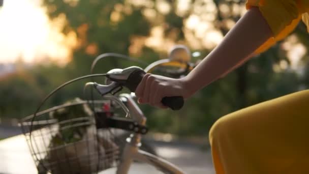 Wyświetlić zbliżenie do poznania womans ręce trzyma na kierownicy roweru podczas jazdy na rowerze miasta z koszem i kwiaty. Flary podczas wczesnych handlowych. Słońce wschodzi. Kobieta w sukni żółty — Wideo stockowe