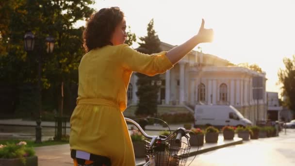 Achteraanzicht van een mooie vrouw een citybike met een mand en bloemen in het centrum van de stad tonen haar duim omhoog rijden. Lens flare. Tikje schot — Stockvideo
