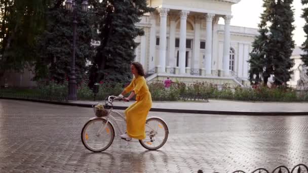 Beautiful woman riding a city bicycle with a basket and flowers in the city center enjoying her time early in the morning. Steadicam shot. Beautiful old city view — Stock Video