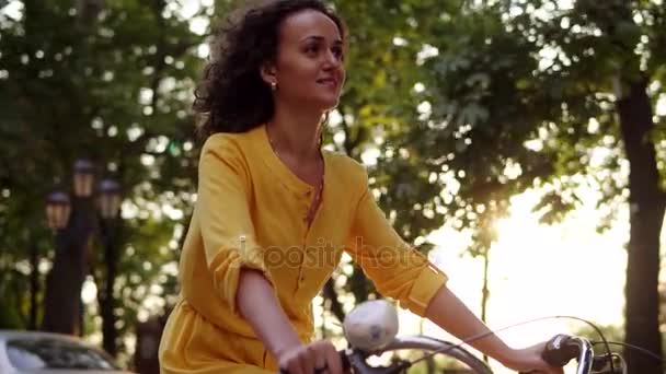 Lens flare: sorridente donna felice in abito lungo giallo è in sella a una bicicletta di città con un cesto e fiori nel parco con alberi verdi intorno durante l'alba. Colpo di rallentamento — Video Stock