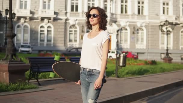 Sorrindo mulher atraente em óculos de sol andando na rua da cidade velha segurando seu longboard na parte da manhã durante o amanhecer. Um tiro em câmara lenta. Bela vista da cidade velha — Vídeo de Stock