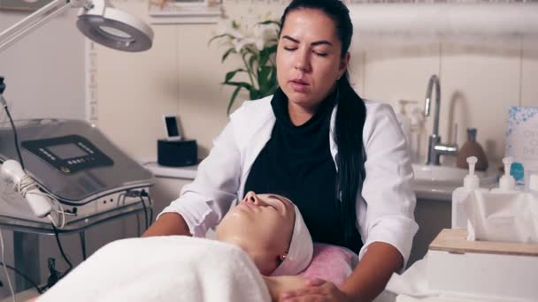 Cosmetologist feminino está fazendo massagem no pescoço no salão de spa. Jovem com os olhos fechados está deitada no sofá durante o procedimento cosmético do rosto e pescoço. Cuidado com o rosto. Conceito de beleza, saúde e juventude — Vídeo de Stock