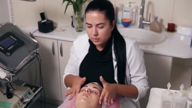 Kadın güzellik uzmanı yüz spa salonda masaj yapıyor. Gözleri kapalı ile genç kadın kozmetik yüz ve boyun yordamı sırasında kanepede yatıyor. Güzellik, sağlıklı ve genç kavramı