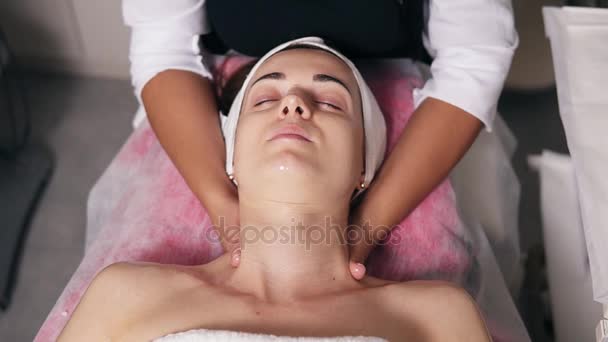 Young woman with her eyes closed is lying on the couch during cosmetic face procedure. Professional cosmetologist is making neck massage in spa salon. Face care. Beauty, healthy and youth concept — Stock Video