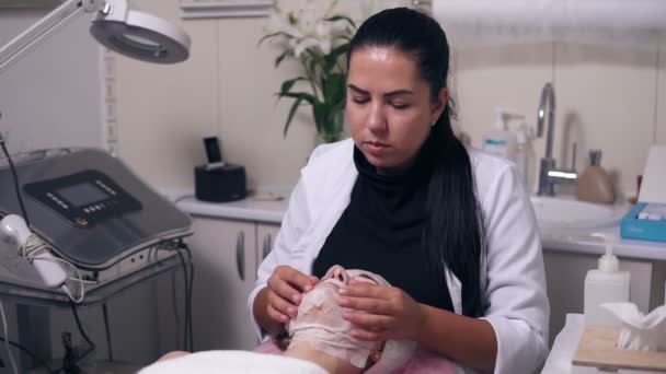 Професійні косметологи руки роблять масаж обличчя молодій жінці з маскою на обличчі та шиї під час косметичної процедури обличчя. Догляд за обличчям — стокове відео
