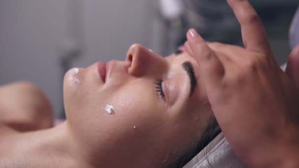 Detailní pohled na ženu s očima zavřené ležet na gauči, zatímco profesionální kosmetička je použitím krém na obličej a obličejem, masáže ve spa salonu — Stock video