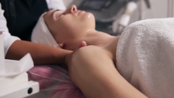 Wyświetlić zbliżenie kobieta z jej oczy zamknięte, leżąc na kanapie podczas procedury kosmetyczne twarzy. Profesjonalne kosmetyczki jest Dokonywanie szyi masaż w salonie spa. Piękna, zdrowa i koncepcja młodzieży — Wideo stockowe