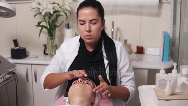 Επαγγελματική cosmetologist είναι κάνοντας μασάζ σε κομμωτήριο spa προσώπου. Νεαρή γυναίκα μιλά της cosmetologist ξαπλωμένοι στον καναπέ κατά τη διαδικασία καλλυντικά προσώπου. 4k πυροβόλησε — Αρχείο Βίντεο