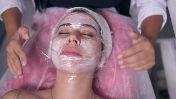 Closeup view of young woman lying on the couch while professional cosmetologist is apllying special treatment on womans face and covers it with polyethylene foil. Professional skin care in spa salon — Stock Video