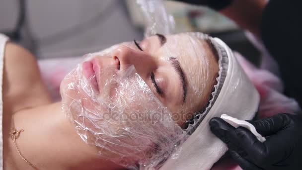 Profesjonalne kosmetyczki jest czyszczenie twarzy womans ze szczególnego traktowania przy użyciu gąbki bawełny. Młoda kobieta leży na kanapie podczas procedury kosmetyczne twarzy w salonie spa — Wideo stockowe
