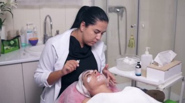 Profesyonel Cilt Bakımı spa salonda genç kadın için. Profesyonel güzellik uzmanı apllying özel muamele kadının yüzünde ise genç kadın kanepede yatıyor