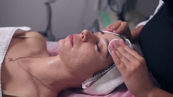 Vista de cerca del cosmetólogo profesional es la limpieza de la cara de las mujeres usando esponja de algodón. Mujer joven está acostada en el sofá durante el procedimiento cosmético de la cara en el salón de spa. Cuidado facial — Vídeos de Stock