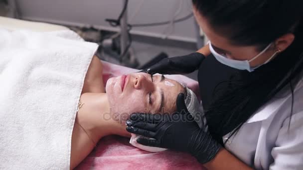 Giovane donna con pelle difettosa sdraiata sul divano mentre il cosmetologo professionista applica la crema per il viso e fa massaggi al viso nel salone spa — Video Stock