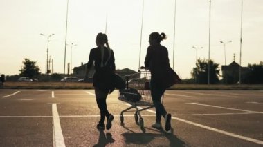 Arkadan görünüşü iki çekici şık kız alışveriş merkezinin park yerinde yürümek. Bir tanesi bir longboard onun elinde tutuyor. Birlikte vakit güzel arkadaşlar. Slowmotion atış