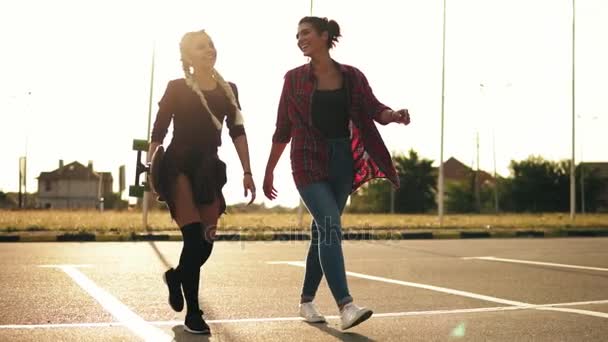 Счастливые стильные девушки-хипстеры гуляют вместе на парковке. Одна девушка с длинными косичками держит скейтборд в руках. Медленный выстрел. Вспышка объектива — стоковое видео