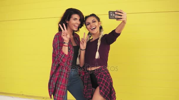 Duas meninas hipster fazendo selfie e mostrando sinal de paz em pé junto à parede amarela. Meninas bonitas posando e tirando fotos com telefone inteligente. Tiro em câmara lenta — Vídeo de Stock