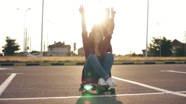 Eine fröhliche Frau sitzt auf einem Longboard und hebt fröhlich die Hände, während ihr Freund sie bei Sonnenuntergang hinter sich herschiebt. Linsenschlag. Zeitlupenschuss — Stockvideo