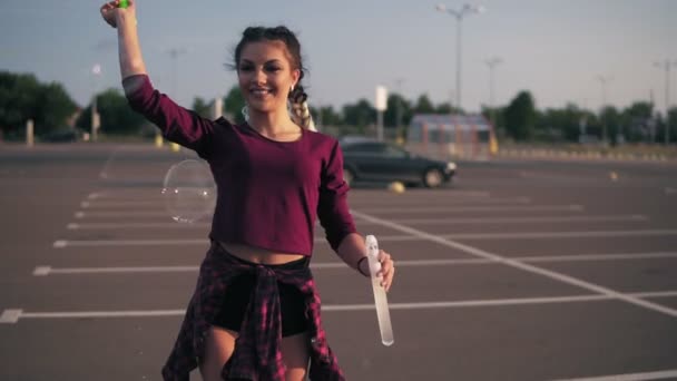 Jovem menina hipster brincalhão com tranças longas soprando bolhas no estacionamento junto ao shopping da cidade durante o pôr do sol — Vídeo de Stock