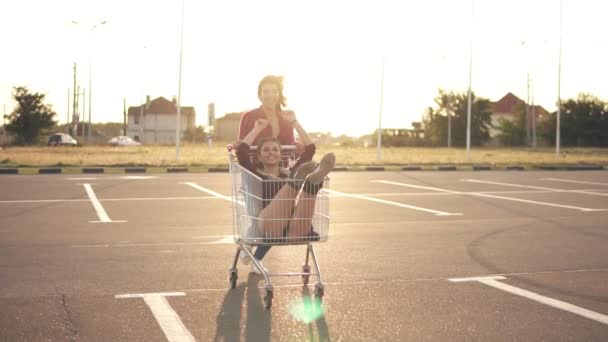 La jeune femme est assise dans le chariot d'épicerie, tandis que son amie la pousse derrière dans le parking près du centre commercial, profitant de l'extérieur avec course de chariot pendant le coucher du soleil. Éclairage des lentilles — Video