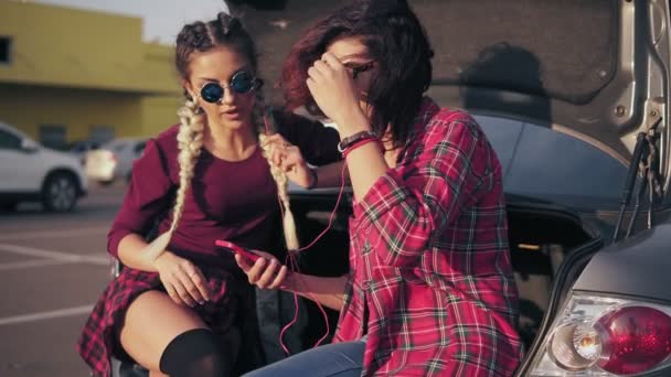 Duas jovens namoradas atraentes em óculos de sol sentadas dentro do porta-malas do carro aberto no estacionamento perto do shopping e ouvindo a música no smartphone. Tiro em câmara lenta — Vídeo de Stock