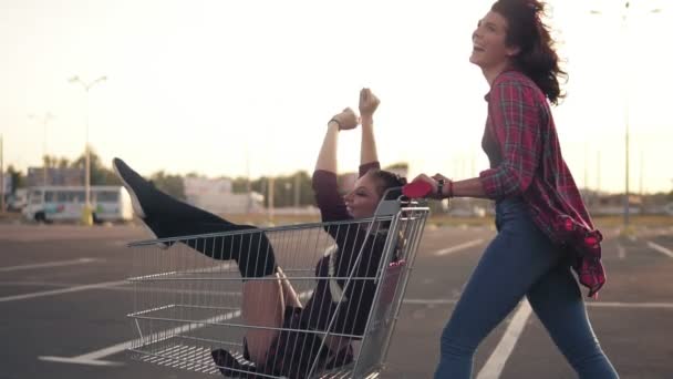 Vista lateral de uma jovem mulher feliz empurrando um carrinho de supermercado com sua namorada dentro no estacionamento junto ao shopping durante o pôr do sol. Tiro em câmara lenta — Vídeo de Stock