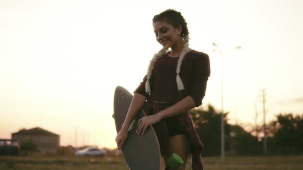 Ung attraktiv kvinna i rutig skjorta, shorts och linne holding longboard och tittar i kameran under solnedgången sommartid. Bländning. Slowmotion skott — Stockvideo