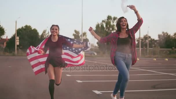 Νέοι ευτυχισμένη αμερικανική hipster κορίτσια τρέχουν ενώ κρατάτε την αμερικανική σημαία και την αφήνει ανοικτά χειροβομβίδα καπνού βόμβα με λευκό χρώμα διασκεδάζοντας. Κοιτάζοντας την κάμερα. Slowmotion βολή — Αρχείο Βίντεο