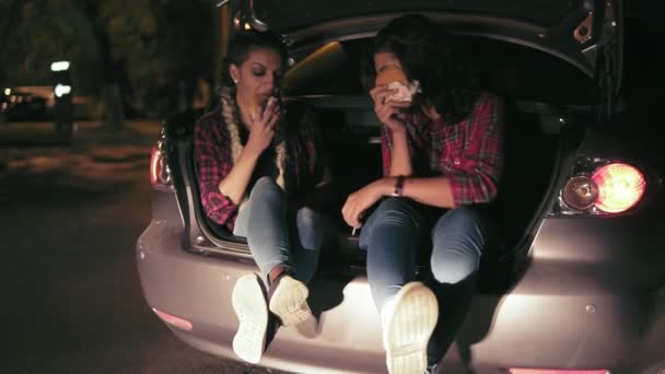 Two attractive girlfriends sitting in the open trunk of the car, talking and eating sandwiches, drinking from the paper cup during the night in the city. Slowmotion shot — Stock Video