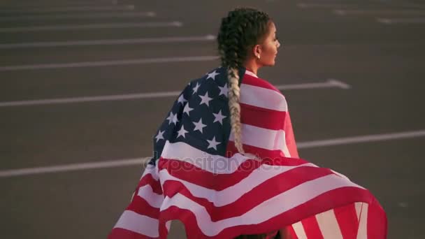 Menina americana orgulhosa segurando estrelas e listras. Ela envolve a bandeira americana sobre seus ombros e caminha para a frente. Vista traseira. Tiro em câmara lenta — Vídeo de Stock